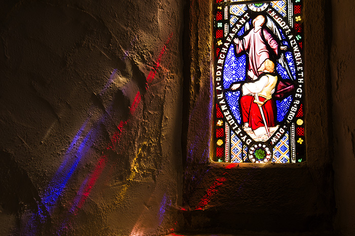 Stained glass window in an ancient church lit from outside, with blue and red shafts of light extending inside onto the stone walls