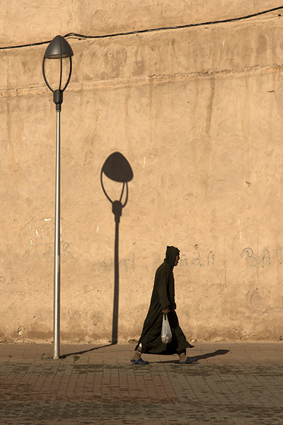 A man in tradition Moroccan clothing walks past a lamp post and it's shadow, carrying a bag of fish