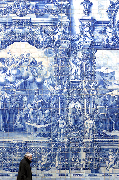 A forlorn looking man walking in front of a giant wall full of blue and white tiles depicting a religious scene