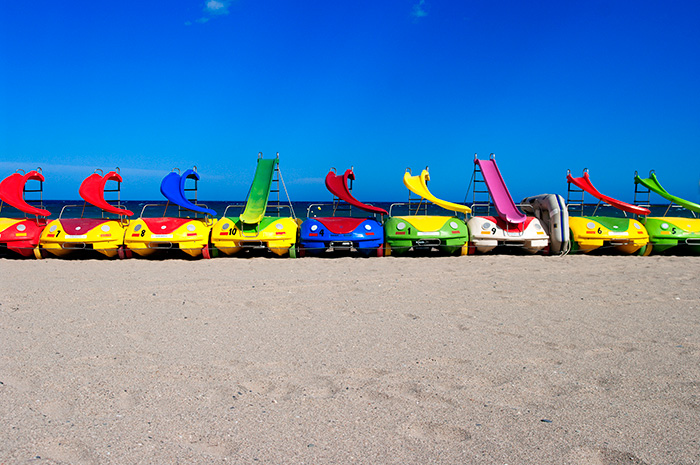 A line of colourful plastic water cars with slides sit on a sandy beach against a deep blue sky