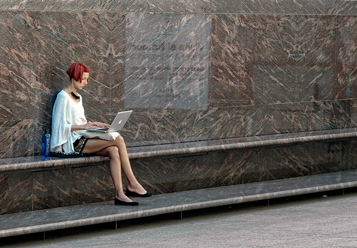 Elegant looking woman with red hair sits against a marble wall with a laptop. The screen reflects onto the wall beside her.