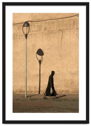 Framed image of a man in tradition Moroccan clothing walking past a lamp post and it's shadow, carrying a bag of fish