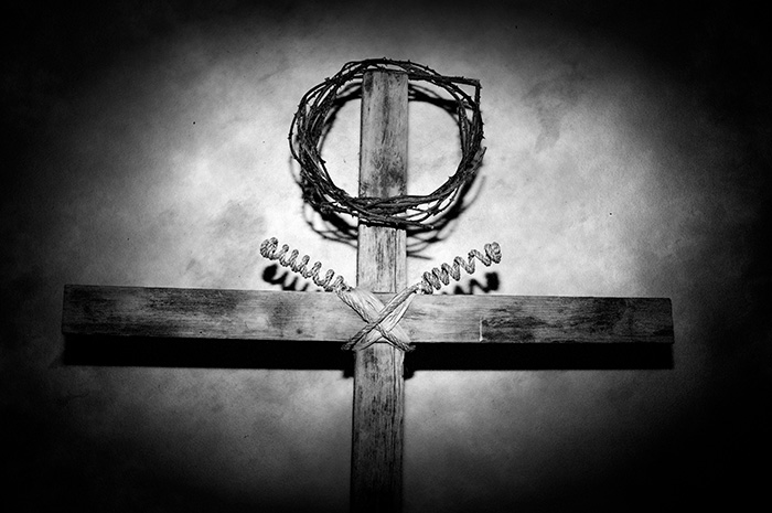 Artistic rendition of a cross with crown of thorns balanced on top leaning against the bare wall of an old church