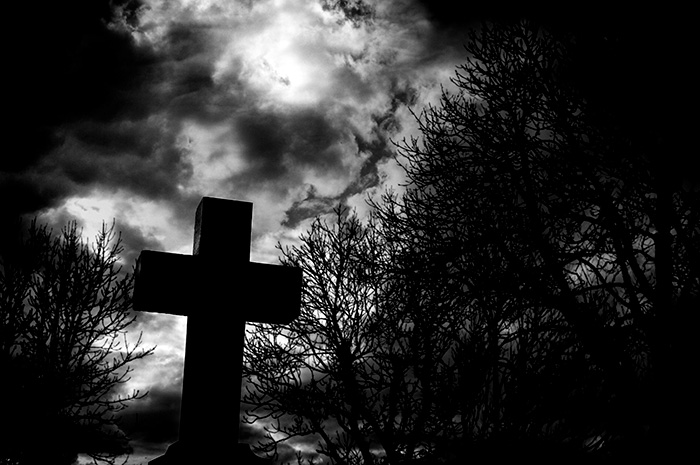 A stone graveyard cross photographed from below with bare trees and a menacing sky in the background