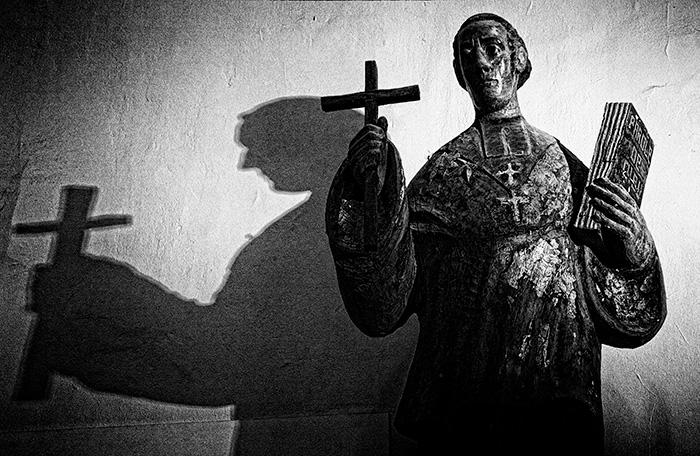 A statue of a preacher holds a cross in one hand and a Bible in the other with a shadow on the wall behind