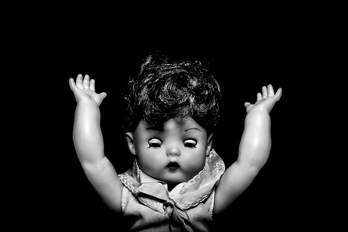 A vintage doll, eyes closed, holding arms in the air, surrounded by blackness