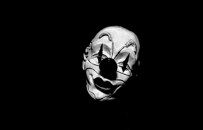 A slightly lopsided clown's face peers out from a black background