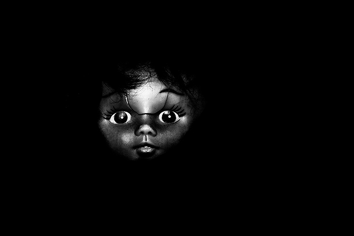Portrait of a grotesque looking doll with a split in her head looking intently forward, surrounded by blackness