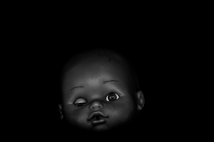 Portrait of grotesque doll with one eye open and one eye closed surrounded by blackness