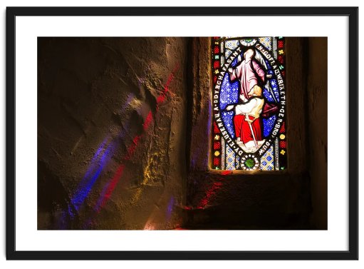 Framed image of a stained glass window in an ancient church lit from outside, with blue and red shafts of light extending inside onto the stone walls